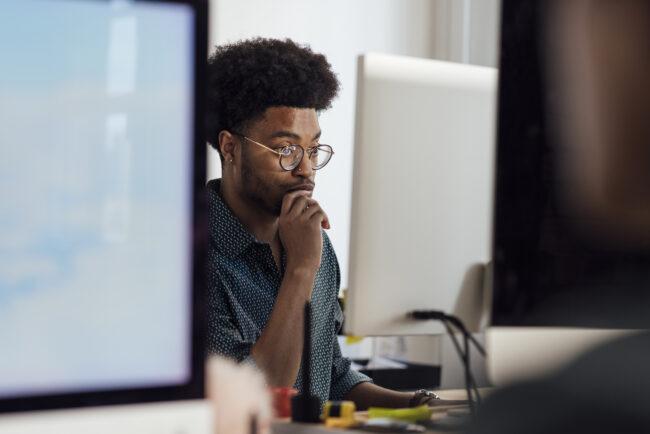 Close-up of a young male office worker sitting at her desk using a computer to complete her work in the office in Newcastle, England. He is wearing smart casual work clothing and he is working to a deadline.