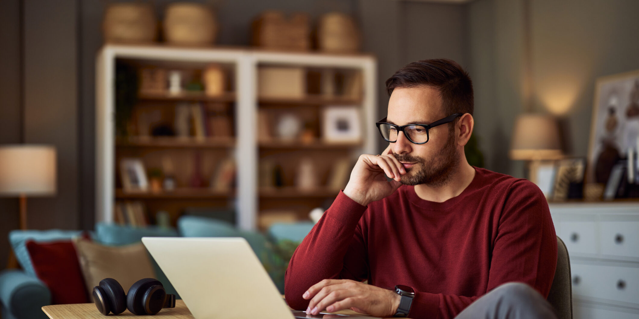 A busy, focused young adult male freelancer sitting at a desk and resting his head on his hand while working on a project on his laptop.